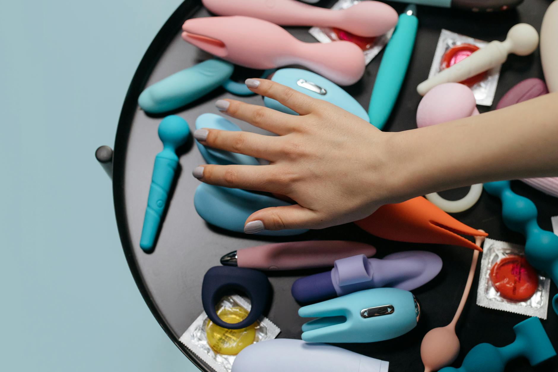 A hand reaching over a variety of colorful intimate wellness devices arranged on a table.