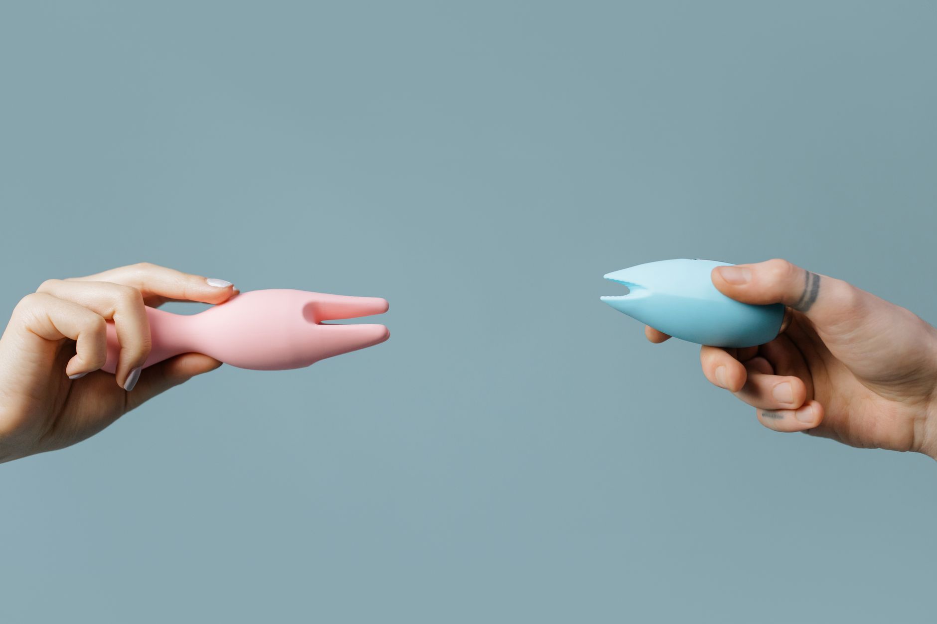 Two hands holding silicone vibrators against a pastel background, representing connection and shared pleasure.
