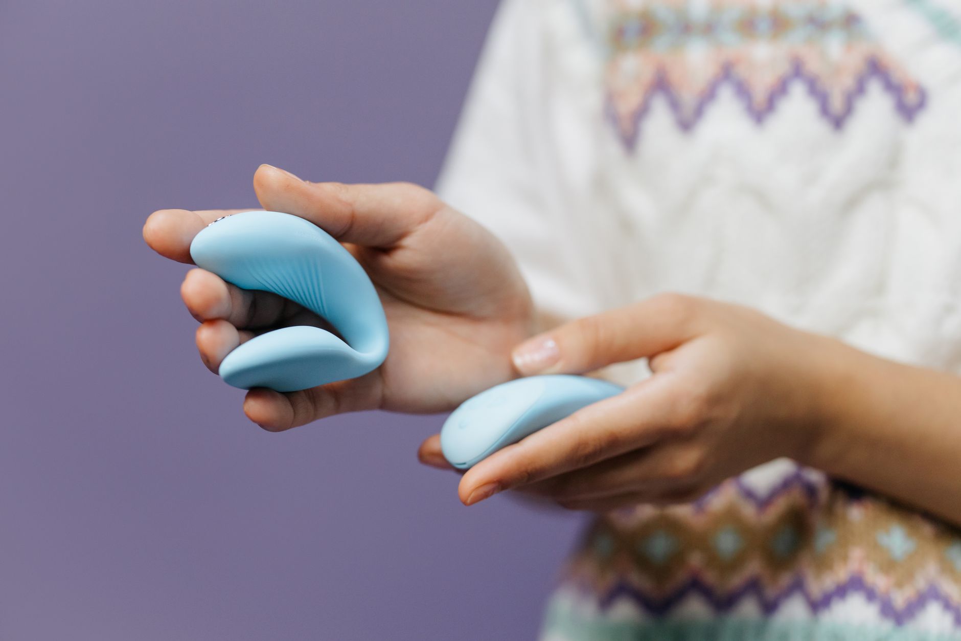 Nahaufnahme von Händen, die einen eleganten blauen Vibrator vor violettem Hintergrund halten.